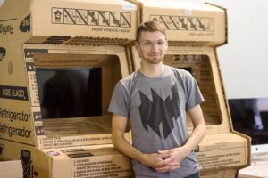 Justin Gitlin stands with cardboard arcade games from OhHeckYeah installations.