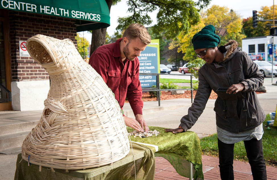 A woman and a man stand at a table with a green tablecloth, a variety of shells, and a woven basket sculpture.