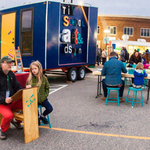 People sit at outdoor tables with art supplies in front of a blue trailer that says Tiny School of Arts & Design