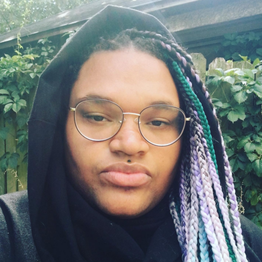 Adrienne Doyle headshot – a Black woman with glasses and a lip piercing, with teal and lilac braids, wearing a black hoodie